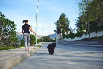 Rear view woman walking her dog on leash in the nature. Smiling female in sportswear, enjoying walk...