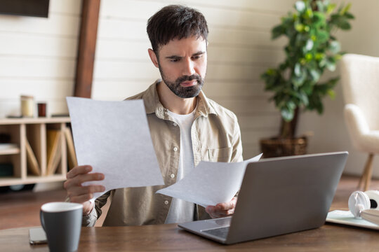 A man with a beard sits at desk intently examining sheets of paper in a well-lit room. In front of him is an open laptop, suggesting he is either working from home or managing personal affairs