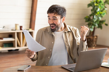 A man in casual attire sits in a home office, expressing surprise and joy while looking at a...