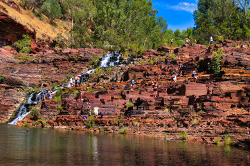 Dozens of people relaxing on the red rocks at Fortescue Falls and in the cool pool at the foot of the falls. Dales Gorge, in the remote outback of Karijini National Park, Western Australia. 
