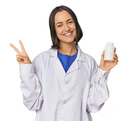 Young Caucasian female pharmacist with pills joyful and carefree showing a peace symbol with...
