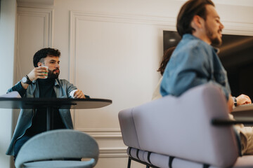 Obraz premium A pensive bearded young man holding a glass while sitting across from a colleague in a relaxed business setting.