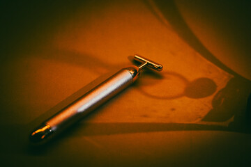 A golden electrical facial T-massager on abstract dark yellow background flatly. Beauty care treatment anti-aging product mock-up. Female routine tool