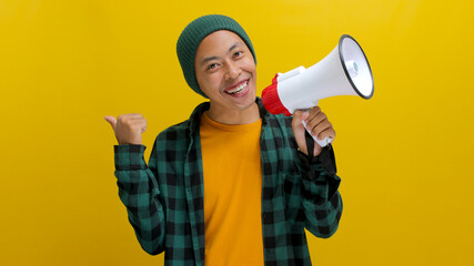 Happy Asian man in a beanie hat and casual shirt talks into a megaphone and points at empty copy...