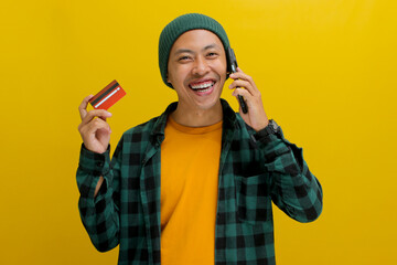 Happy young Asian man, wearing beanie hat and casual shirt, holds a credit card while engaging in a phone call, confirming a purchase via telephone with customer service, isolated on yellow background