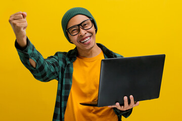 Happy young Asian man, dressed in a beanie hat and casual shirt, pumps his fist in excitement while holding a laptop, expressing a YES gesture, celebrating career growth and success in business.