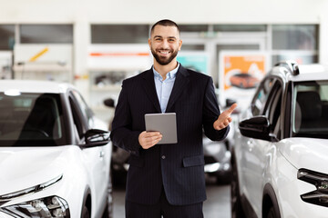A man stands inside a car showroom, holding a tablet and looking at it intently. The showroom is...