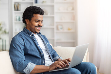 African American man sitting comfortably on a couch, using a laptop computer. He is engrossed in...