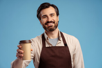 Young barista barman wearing brown apron holding craft paper brown cup of coffee