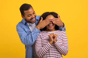 A smiling young African American man is standing behind a woman, playfully covering her eyes with...