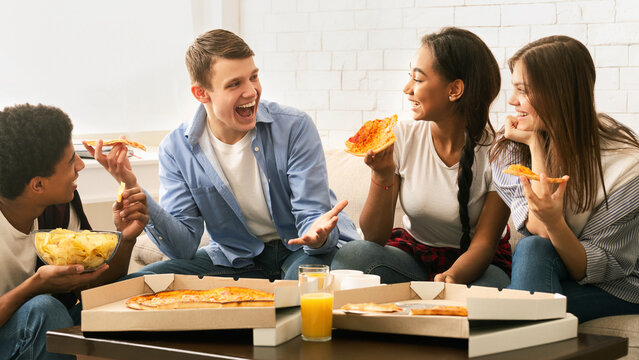 A cheerful group of four young friends is gathered in a comfortable living room, enjoying slices of pizza and engaging in a lively conversation