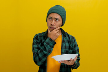 Pensive young Asian man, dressed in a beanie hat and casual shirt, ponders over what to eat while holding an empty white plate, contemplating his menu for cooking