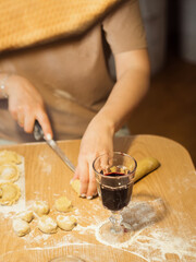 A vertical close-up of a woman making dumplings, her face obscured by a straw lampshade, as she skillfully cuts dough with a knife. Focus on the glass of red wine in the foreground..