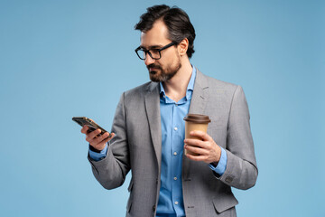 Young bearded man in jacket and glasses reading message on phone, holding coffee in studio