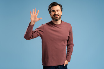 Smiling bearded man in casual waving by hand while posing in studio, looking at camera