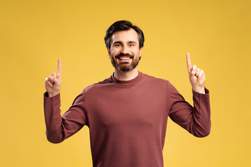 Smiling bearded young man pointing up by fingers posing in studio, isolated on yellow