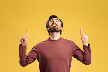 Happy bearded young man looking up and pointing by fingers posing in studio, isolated on yellow