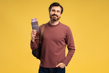 Smiling man holding passport with plane ticket and backpack on yellow background