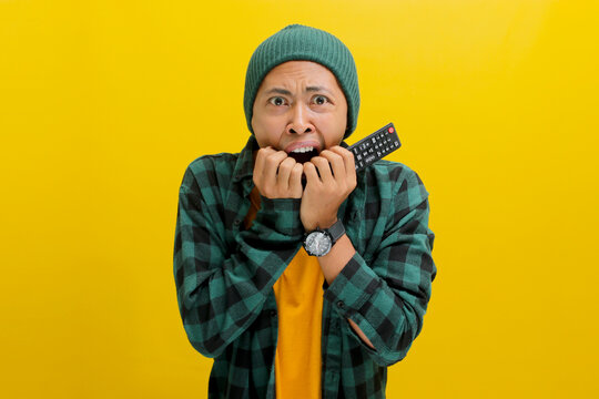 Scared Asian man, adorned in beanie hat and casual shirt, holds TV remote, visibly startled by a jump scare while watching a horror movie, feeling frightened and fearful, isolated on yellow background