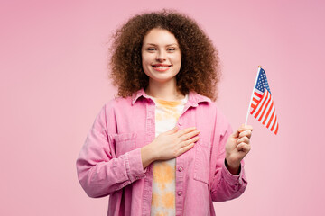 Beautiful, happy young woman, patriot holding American flag with hand on heart, looking at camera