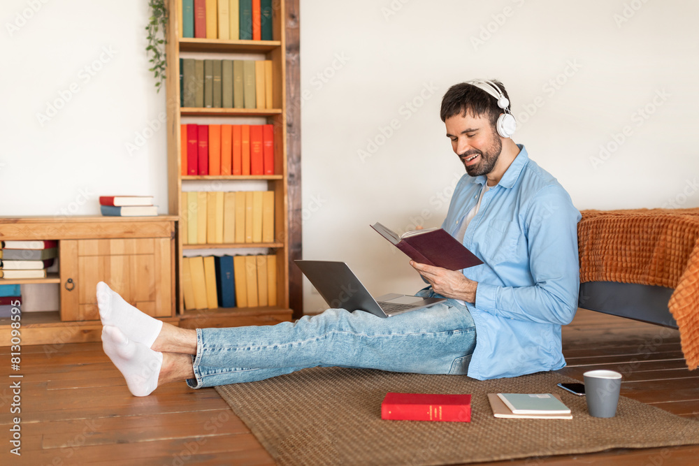 Poster A man is seated on the floor, engrossed in a book while typing on a laptop placed in front of him. He appears focused and concentrated on his work, surrounded by a simple and minimalistic setting.
