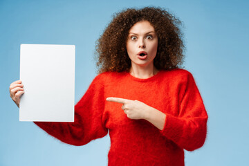 Shocked, curly haired young woman holding white, blank paper pointing finger at mockup