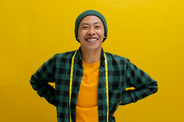 Cheerful Asian man in a beanie and casual shirt smiles at the camera, holding a yellow measuring tape around his neck. Isolated on a yellow background