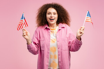 Overjoyed woman holding American flag, looking at camera, standing isolated on pink background