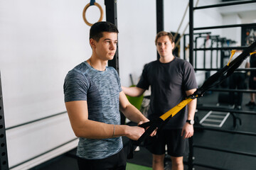 Skilled fitness instructor male showing suspension training to motivated beginner young man, performing pull-ups exercise with trx straps at gym. Lifting up and down to maintaining muscles.