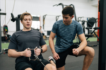 Portrait of beginner sportsman training in gym with personal trainer, explaining how to work on rowing machine. Man doing exercises for muscles under supervision of coach measuring time on stopwatch