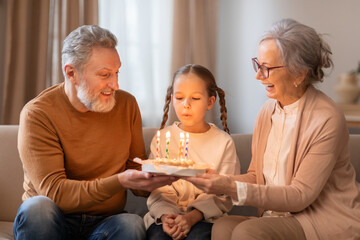 A young girl sitting between her smiling grandparents is blowing out candles on a birthday cake...