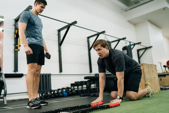 Exhausted beginner sportsman falling to floor from fatigue after battle rope workout in cross training gym with help of personal trainer. Concept of healthy lifestyle, physical sport activity.