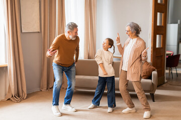 An elderly couple is joyfully dancing with a young child in the warmth of a well-lit living room....