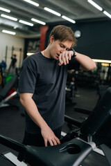 Vertical portrait of tired runner male resting after running on treadmill during cardio session in gym. Satisfied fit young man standing on treadmill in fitness gym, feeling exhausted after jogging.