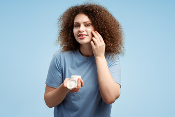 Happy attractive young woman with curly hair applying cream on face, looking in mirror, standing