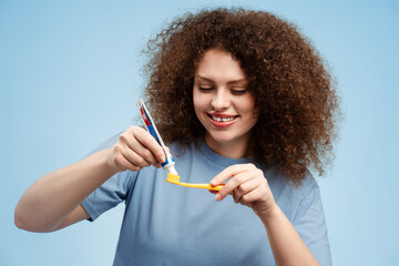 Beautiful, smiling young woman with curly hair holding toothbrush and toothpaste, brushing her teeth