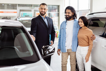 A young Indian couple is smiling and standing next to a new car at a dealership, interacting with a professional salesman who is offering the car keys - Powered by Adobe