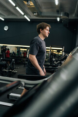 Vertical shot of beginner sportsman having intensive cardio workout to burn calories in gym. Side view of young athletic man jogging on gym machine, having cardio session in well-equipped fitness club