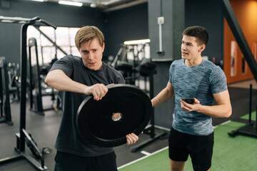 Portrait of motivated beginner sportsman hard time spinning barbell disks overhead at fitness gym support under supervision of personal trainer. Concept of healthy lifestyle, physical sport activity.