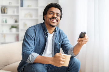 African American man is seated on a couch, holding a remote control in his hand. He appears to be...