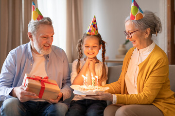 A young girl adorned with a birthday hat is sitting between her grandparents, who are smiling...