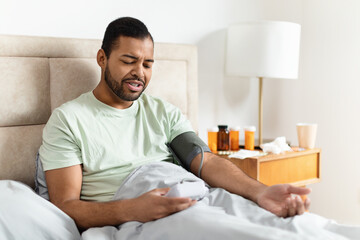 A young African American man is sitting up in bed, wearing a casual T-shirt and using a digital blood pressure monitor on his arm