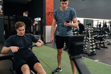 Young tired beginner sportsman training in gym with personal trainer, explaining how to work on rowing machine. Man doing exercises for muscles under supervision of trainer measuring time on stopwatch