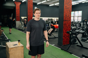 Portrait of beginner athlete male in sportswear smiling looking away standing posing in gym. Determined young man in activewear at sport club. Sportsman after intense crossing training workout session