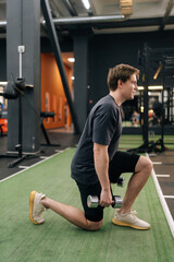 Vertical side view of motivated young beginner athletic sportsman in activewear holding dumbbells doing squat lunges warm up training indoor at gym. Concept of healthy lifestyle, physical activity.