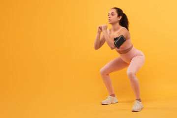 A young woman, dressed in activewear, is captured mid-motion as she performs a squat exercise, focused and maintains good form with her arms held in front of her, workout against yellow background