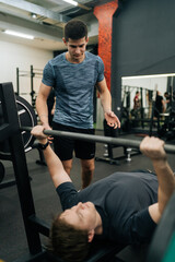 Vertical shot of focused beginner male athlete performing incline bench press under watchful eye of personal trainer at gym. Athletic young man pumping up chest muscles during hard training.