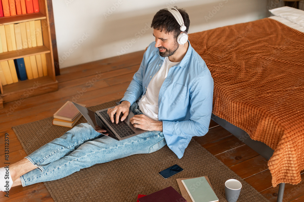 Poster A man is sitting on the floor indoors, focused on using a laptop. He appears to be working or browsing the internet, with the laptop open in front of him, high angle view