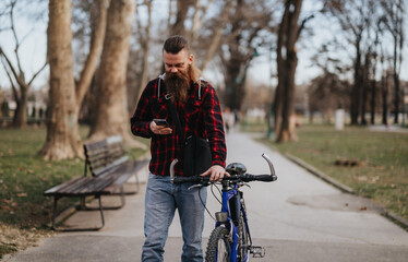 Obraz premium A stylish businessman with a beard, using a smart phone while standing next to his bicycle in an urban park area.