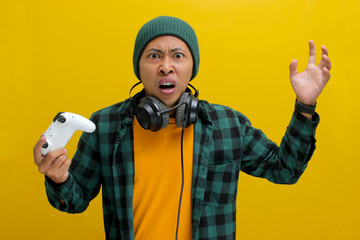 An angry and displeased young Asian man, dressed in a beanie hat and casual shirt, appears stressed while playing a video game using a gamepad on a game console, isolated on yellow background